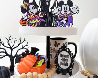 May include: A two-tiered Halloween-themed tray with Mickey and Minnie Mouse cutouts in costume. The tray includes an orange pumpkin candy dish, a mug, and a sign that reads "Welcome Foolish Mortals". Decorative wooden beads and a black tree are also included.