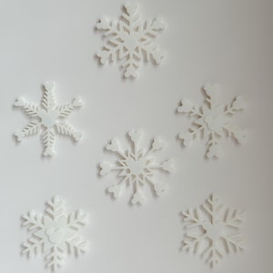 May include: Six white snowflake decorations of varying designs are arranged on a white background. Each snowflake has intricate cut-out patterns, creating a delicate and festive appearance. The snowflakes are ideal for winter or holiday decor.