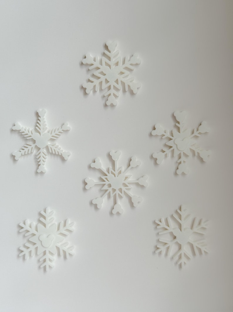 May include: Six white snowflake decorations of varying designs are arranged on a white background. Each snowflake has intricate cut-out patterns, creating a delicate and festive appearance. The snowflakes are ideal for winter or holiday decor.