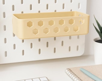 May include: A rectangular, pale yellow plastic storage bin featuring a honeycomb-patterned cutout design on the front panel. The bin is mounted onto a white pegboard wall organiser, providing a functional storage solution for small items.