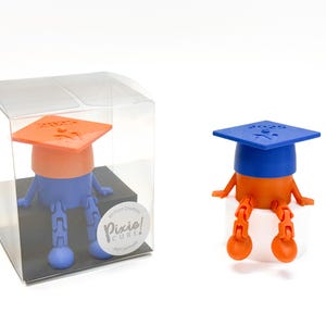 May include: A graduation-themed figurine in orange, blue, and white. The figurine is wearing a graduation cap with the year 2023 on it. The figurine is sitting down with its legs extended. The figurine is in a clear plastic box.