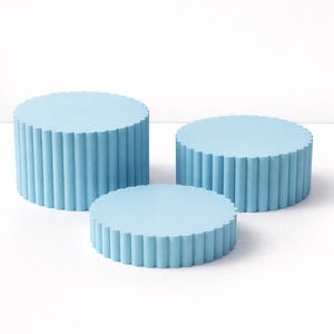 May include: Three light blue, cylindrical display stands with a fluted edge design. The stands vary in height and diameter, creating a tiered effect. The surfaces are smooth and the color is a soft, pastel blue. These decorative stands are ideal for showcasing products.