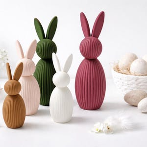 Pu&ograve; includere: Una collezione di figurine decorative a forma di coniglietto pasquale in vari colori, tra cui marrone, rosa, verde, bianco e bordeaux. I coniglietti a coste sono disposti con un cestino di uova maculate e fiori decorativi.