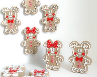 May include: A collection of gingerbread-style ornaments in the shape of a cartoon mouse. Some have red bowties, others have red polka-dot bows. The ornaments are tan with white icing details and red accents.