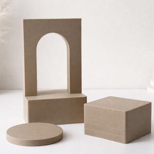 May include: Neutral-toned display props, including a square box, a circular platform, and an arched structure. A white vase with dried pampas grass and a stack of books are in the background. The props are designed for product photography.