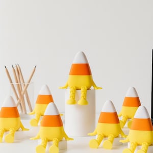 Può includere: Una collezione di figurine fantasiose a forma di caramelle di mais con colori bianco, arancione e giallo. Ogni figurina ha braccia e gambe, conferendo loro un aspetto giocoso. Le figurine sono disposte su una superficie bianca, suggerendo un tema di Halloween o stagionale.