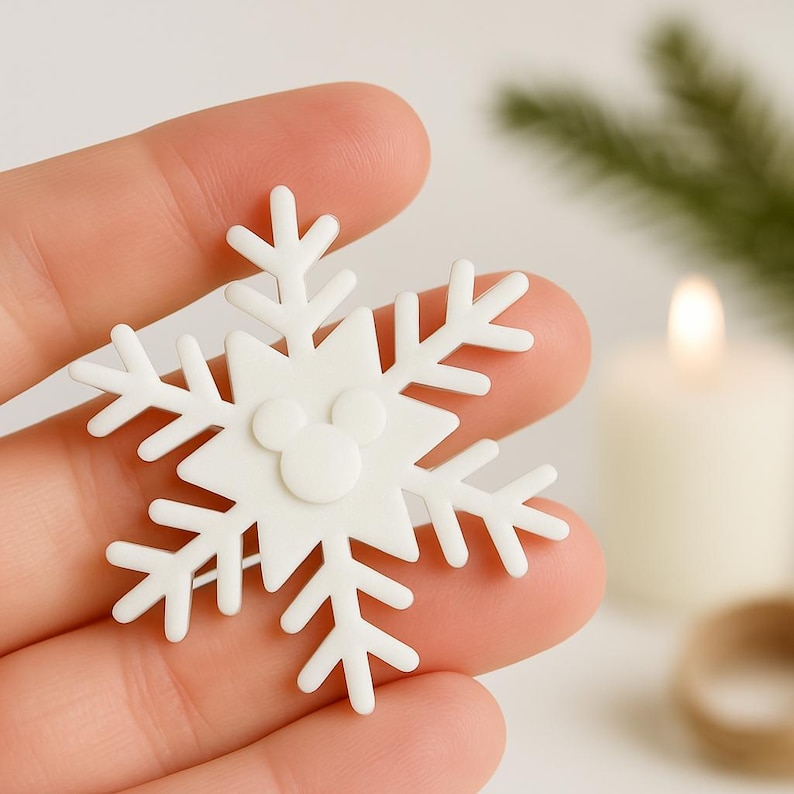 May include: A white snowflake-shaped ornament with a Mickey Mouse head in the center. The ornament has multiple points and is held in a hand. The background includes a lit candle and greenery.