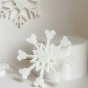 May include: White snowflake decorations with a Mickey Mouse head design at the end of each point. The snowflakes are on a white background with a cylindrical object in the background. The snowflakes are a winter holiday theme.