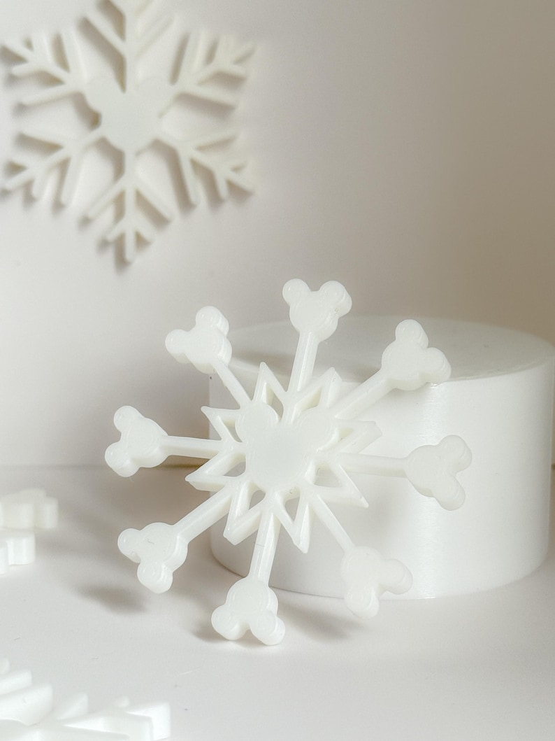 May include: White snowflake decorations with a Mickey Mouse head design at the end of each point. The snowflakes are on a white background with a cylindrical object in the background. The snowflakes are a winter holiday theme.