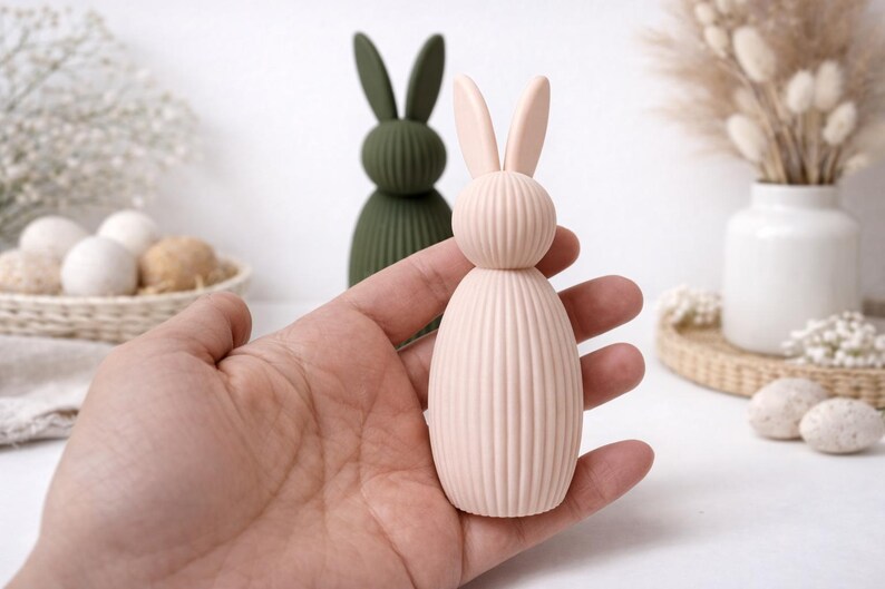 Pu&ograve; includere: Una statuetta di coniglio rosa chiaro e a coste con orecchie dritte &egrave; tenuta in mano. Un'altra statuetta di coniglio verde &egrave; sullo sfondo. Le statuette sono decorative e a tema pasquale.