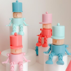 May include: Four colorful toy soldier figures in shades of pink, red, and blue, each with a cylindrical hat and articulated limbs. They are posed sitting on white cylindrical platforms.