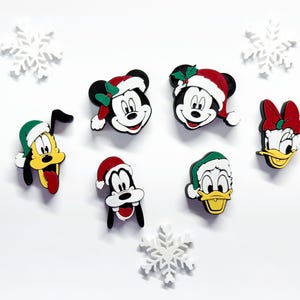May include: A collection of Disney character face ornaments, including Mickey Mouse, Minnie Mouse, Pluto, Goofy, and Donald Duck, all wearing Santa hats. White snowflake decorations are scattered around the characters.