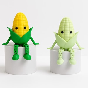 May include: Two articulated corn figurines with black eyes and green husks. One figure is bright yellow with dark green husks and legs, while the other is a pale light green throughout. Both figures have jointed limbs and sit upright.