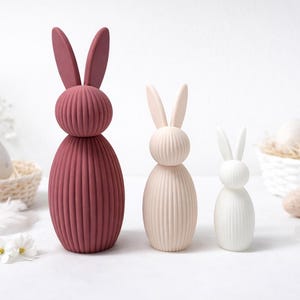 Pu&ograve; includere: Tre statuette decorative a forma di coniglietto pasquale di varie dimensioni e colori. Il pi&ugrave; grande &egrave; di colore rosa intenso, quello centrale &egrave; beige chiaro e il pi&ugrave; piccolo &egrave; bianco. Ognuno ha un corpo a coste e orecchie lunghe. Uova di Pasqua e fiori sullo sfondo.