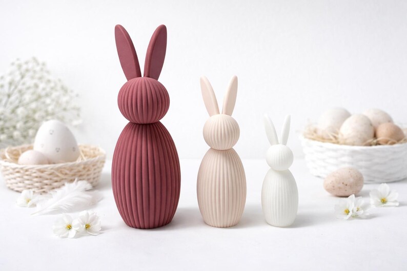 Pu&ograve; includere: Tre statuette decorative a forma di coniglietto pasquale di varie dimensioni e colori. Il pi&ugrave; grande &egrave; di colore rosa intenso, quello centrale &egrave; beige chiaro e il pi&ugrave; piccolo &egrave; bianco. Ognuno ha un corpo a coste e orecchie lunghe. Uova di Pasqua e fiori sullo sfondo.
