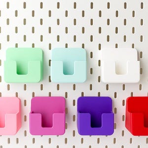 Puede incluir: Siete organizadores de pared cuadrados y coloridos en tonos verdes, azules, blancos, rosas, morados y rojos. Cada organizador tiene un bolsillo en forma de U. Están montados en una pared perforada blanca con agujeros espaciados.
