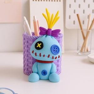 May include: A whimsical blue desk organizer shaped like a cartoon character, with yellow hair and a purple bow. The organizer has a purple woven basket design and holds pens. The character has a red collar and stitched details.