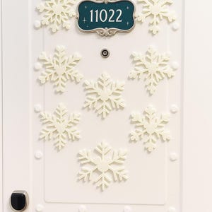 Puede incluir: Puerta blanca decorada con adornos de copos de nieve color crema. Un letrero verde azulado con el número "11022" está centrado encima de la mirilla de la puerta. La puerta tiene un tirador plateado y una cerradura electrónica negra.