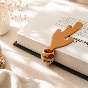 Puede incluir: Un marcapáginas marrón con forma de tarro de miel y un diseño de miel goteando hacia arriba. El tarro tiene la palabra HUNNY impresa en letras negras en la parte frontal. El marcapáginas está colocado dentro de un libro blanco con texto negro.