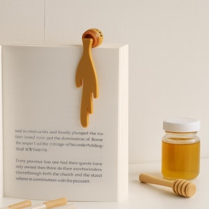 以下が含まれることがあります： 本に付いている、液だれのような形をしたマスタードイエローのブックマーク。ブックマークの上部は丸みを帯びており、数字の「2」が書かれています。小さな蜂蜜の瓶と木製のハニーディッパーも写っています。