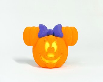 May include: An orange pumpkin-shaped decoration with a smiling face and a purple bow. The pumpkin has two orange ear-like shapes on the sides. The face is lit up with a yellow glow.