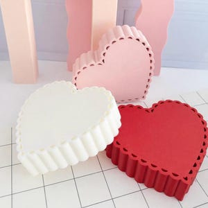 May include: Three heart-shaped objects with scalloped edges and ribbed sides, presented in solid white, soft pink, and deep red colours. Each piece features a distinct, uniform texture along the perimeter and a smooth, flat top surface.