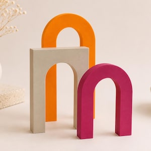 Puede incluir: Tres objetos decorativos en forma de arco de diferentes tamaños y colores. El arco más alto es de color naranja brillante, el arco mediano es de color beige claro con un marco rectangular y el más pequeño es de color rosa magenta, todos erguidos.
