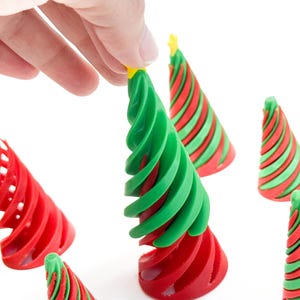 May include: A hand holding a green and red spiral Christmas tree decoration with a yellow star on top. Other similar decorations are scattered around. The decorations are red and green, with a spiral design. The background is white.