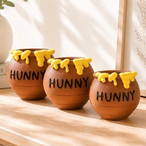 May include: Three brown ceramic pots of varying sizes, each featuring the word HUNNY in black lettering on the front. The rims of the pots are decorated with a yellow, dripping honey-like design that extends down the upper exterior surface.