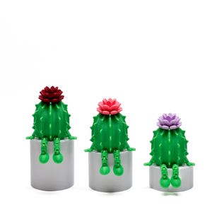 Puede incluir: Tres figuritas de cactus verdes con flores de diferentes colores en la parte superior. Cada cactus está sentado en un pedestal gris.