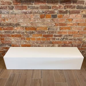 May include: A white rectangular platform or pedestal, likely used for displaying products or as a decorative element. It is sitting on a tiled floor in front of a brick wall.