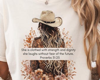 May include: Ivory-colored t-shirt featuring a graphic of a woman in a cowboy hat, surrounded by floral elements. The text "She is clothed with strength and dignity she laughs without fear of the future. Proverbs 31:25" is printed below the graphic.