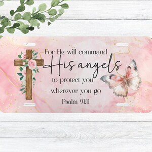 May include: A pink and gold watercolor background with a wooden cross adorned with white and pink flowers. The text "For He will command His angels to protect you wherever you go Psalm 91:11" is written in black script. A pink butterfly with gold accents is on the right side of the image.