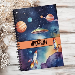 May include: A spiral bound notebook with a space-themed design. The cover features a watercolor illustration of planets, stars, a UFO, and rockets. The name "JACKSON" is printed in black on a peach-colored banner.