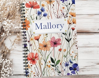 May include: A spiral-bound notebook featuring a white cover with a colorful wildflower pattern in shades of red, orange, pink, and blue. A rectangular white label is centered on the cover with the name Mallory printed in dark blue text.