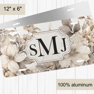 May include: A 12 inch by 6 inch personalized aluminum license plate with a floral design in shades of beige and white. The initials "SMJ" are displayed in a black font within a white frame.