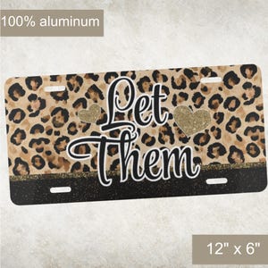 May include: Rectangular license plate with a leopard print background and the words "Let Them" in a cursive font. Two gold glitter hearts are on the plate. The plate is 12" x 6" and made of 100% aluminum.