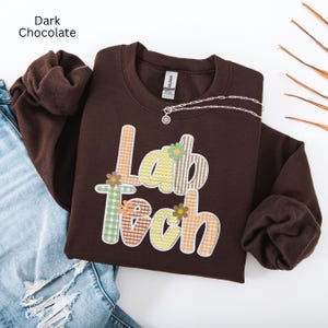 May include: Dark chocolate-colored sweatshirt with the words "Lab Tech" in a colorful, patterned design. The letters are decorated with floral accents. A silver necklace with a pendant is draped across the sweatshirt.