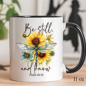 May include: White ceramic mug with a black handle and rim, featuring a watercolor design of a dragonfly, sunflowers, and blue flowers. The mug has the text "Be still, and know" and "Psalm 46:10". The mug is 11 oz.