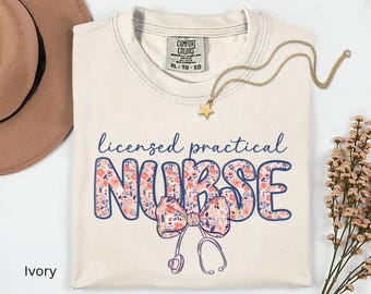 May include: Ivory-colored t-shirt with the words "licensed practical NURSE" in blue script and floral print. A bow and stethoscope graphic are below the text. A gold star necklace and a brown hat are also visible.