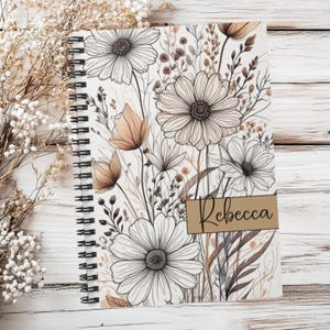 May include: A spiral-bound notebook featuring a white cover with a black line art floral pattern of daisies and wildflowers in shades of tan and brown. A tan rectangular label on the cover contains the name Rebecca in black cursive script.