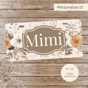 May include: A license plate with the name "Mimi" in a brown frame, surrounded by floral designs in shades of white, brown, and orange. The plate is 12" x 6" and made of 100% aluminum. The text "Personalize it!" is at the top.