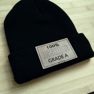 Könnte beinhalten: Schwarze Mütze mit einem braunen Leder-Patch, auf dem "100% ASSHOLE GRADE A" steht.