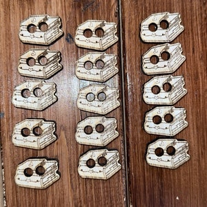 以下が含まれることがあります： 2つの穴のある木製のブック型のボタンが15個セットになっています。ボタンは薄い茶色で、表紙にハートが付いた本の白いデザインが施されています。