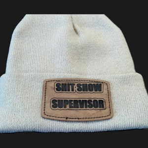 May include: A cream-colored knit beanie with a brown leather patch. The patch features the words "SHIT SHOW" above "SUPERVISOR" in black, bold letters. The hat is a casual accessory.