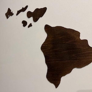 以下が含まれることがあります： ハワイ諸島をモチーフにした木製のウォールアート。島々は濃い茶色の木材から切り出され、白い壁に取り付けられています。島の形が明確に定義されており、州のユニークな地理を示しています。