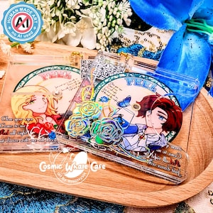 Może przedstawiać: Przezroczyste etui na telefon z akrylu z kolorowymi ilustracjami. Jedno etui przedstawia blond postać, drugie brunetkę. Oba etui mają napis "FEEL" i kwiatowe akcenty. Etui leżą na drewnianej tacy.