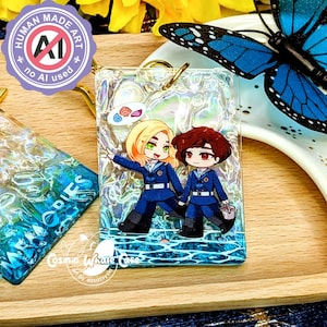 May include: A rectangular resin keychain with two anime-style characters in blue uniforms, holding hands. The keychain has a gold-coloured ring and a holographic background. The text "HUMAN MADE ART" and "no AI used" are visible.