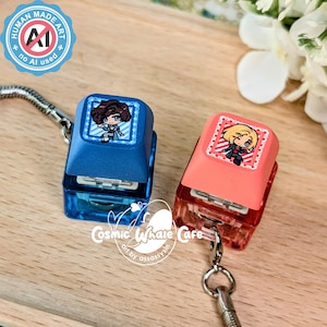 May include: Two mechanical keyboard keycap keychains, one blue and one coral red, each featuring a square graphic of an anime-style character. The blue keycap displays a character with dark hair, while the coral red keycap shows a character with blonde hair.