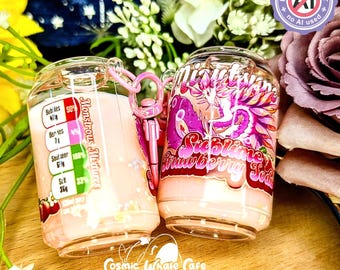 May include: Two clear, soda can-shaped keychains filled with a pink liquid. One keychain reads "Monstrous Mixtures" and the other reads "Sublime Strawberry Soda". Both have pink accents and a pink keychain clasp. The background includes flowers.